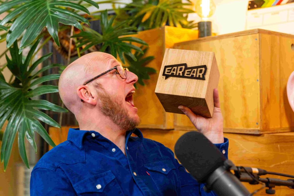 Man in blauwe blouse houdt een houten blok met het logo "EAR EAR" omhoog tijdens een studioshoot.