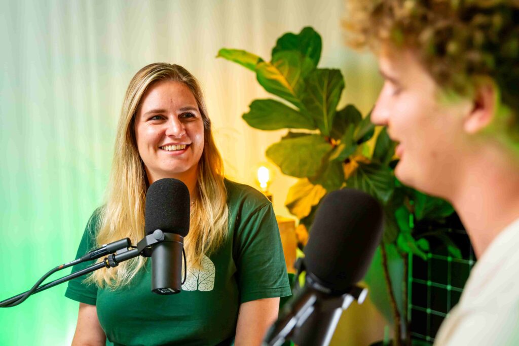 Twee mensen in gesprek tijdens een opname van de Groenpand Podcast, met microfoons en een plant op de achtergrond.