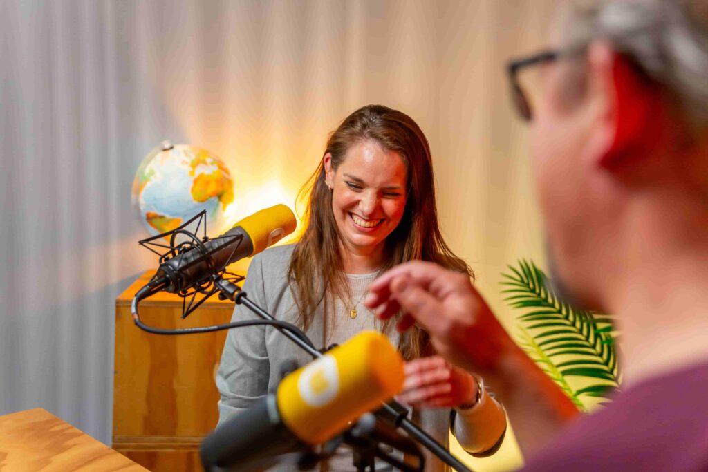 Vrouw lacht tijdens een podcastopname met gele microfoons en wereldbol op de achtergrond.