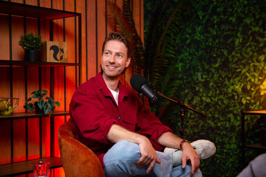 Man in rode blouse spreekt in microfoon tijdens opname van de Groeivoer Podcast in een sfeervolle studio met plantenwand.