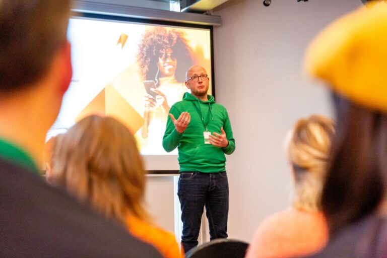 Man in groene trui geeft presentatie tijdens All Ears Club op 17 april 2024, met publiek op de voorgrond.