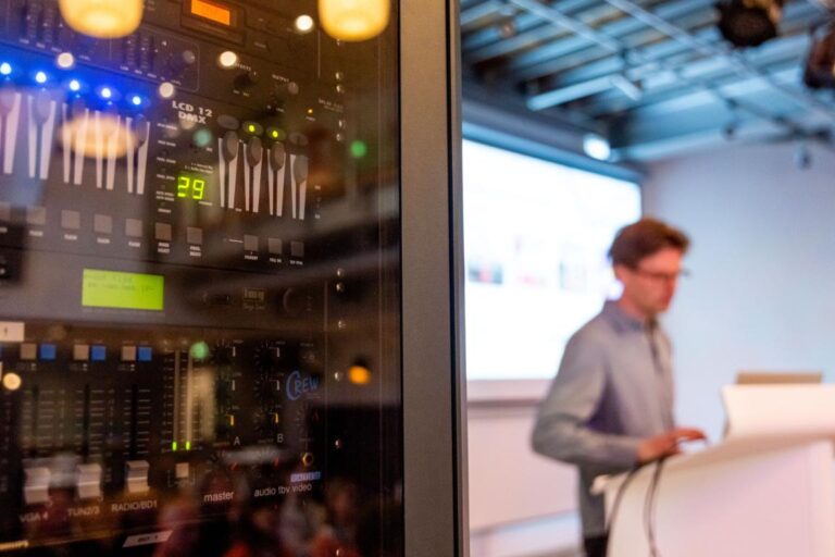 Een presentator bij een All Ears Club-evenement staat onscherp op de achtergrond terwijl scherp in beeld een geluidsinstallatie wordt weergegeven.