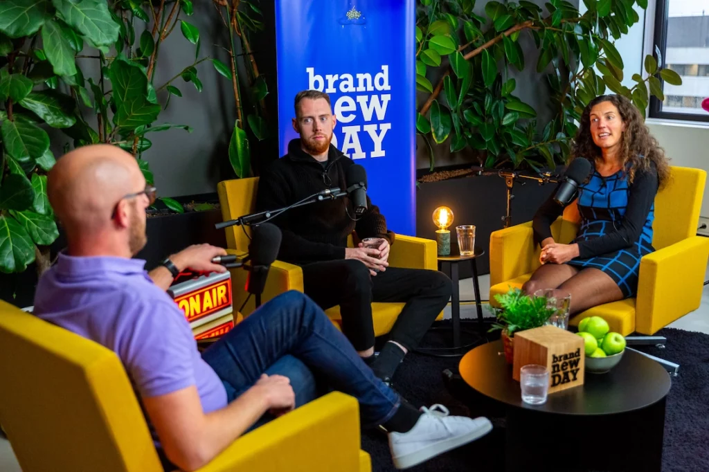 Drie mensen zitten in gele stoelen in een kleurrijke studio en nemen een podcast op voor Brand New Day.