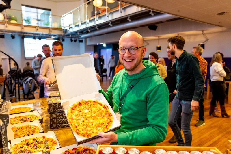 Man in groene trui toont een pizza tijdens een gezellige EarEar-communitybijeenkomst met meerdere mensen en tafels vol eten.