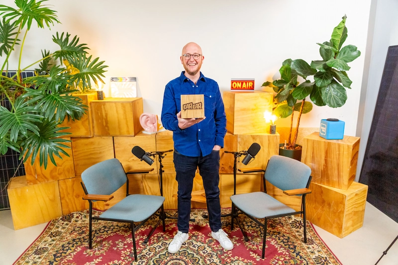 Man staat glimlachend in een kleurrijke podcaststudio met een bordje met het logo "EarEar" in zijn hand.