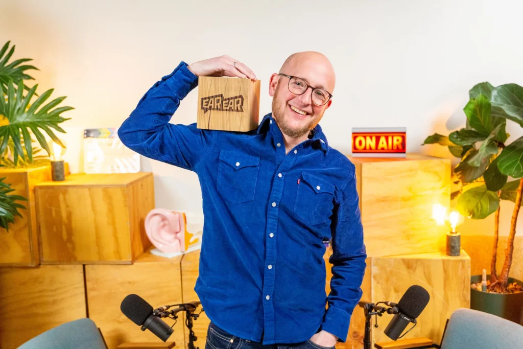 Man in blauwe blouse houdt een houten blok met het logo "EarEar" vast in een kleurrijke podcaststudio.