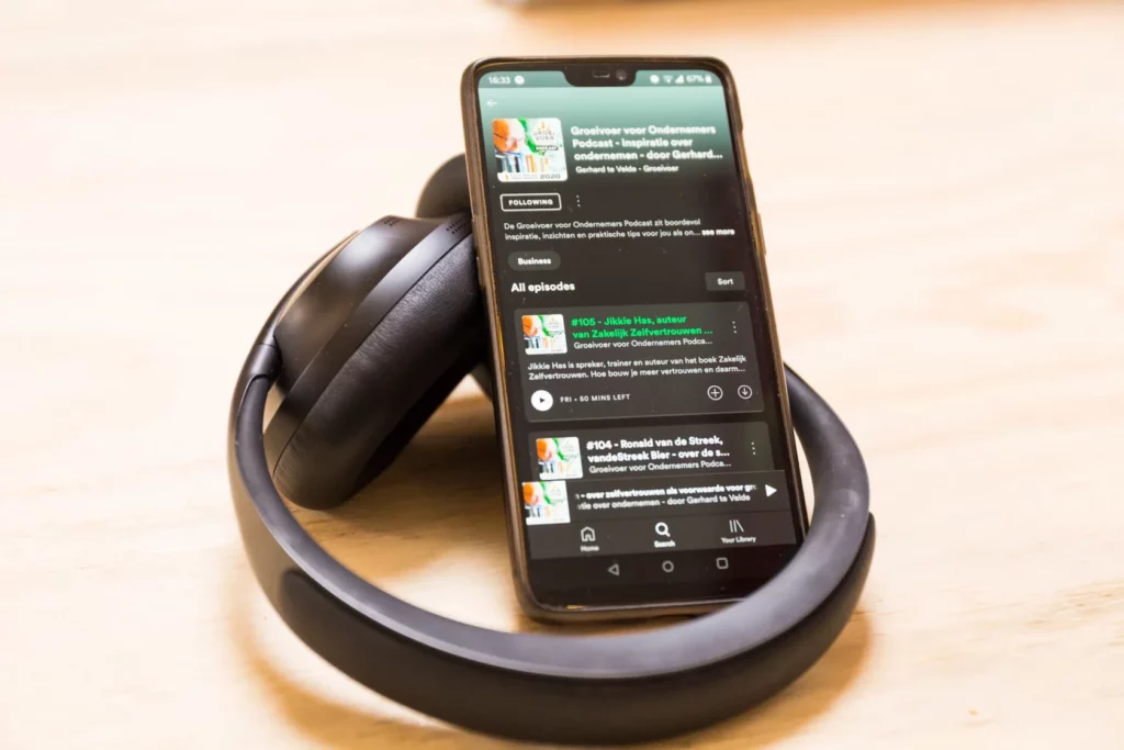 Smartphone met de podcast "Groeivoer voor Ondernemers" op Spotify, omringd door een zwarte koptelefoon op een houten ondergrond.