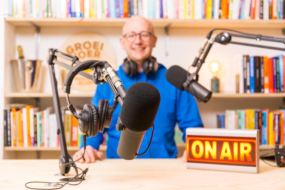 Man in een podcaststudio met microfoons, koptelefoon en een "ON AIR"-bord op de voorgrond.