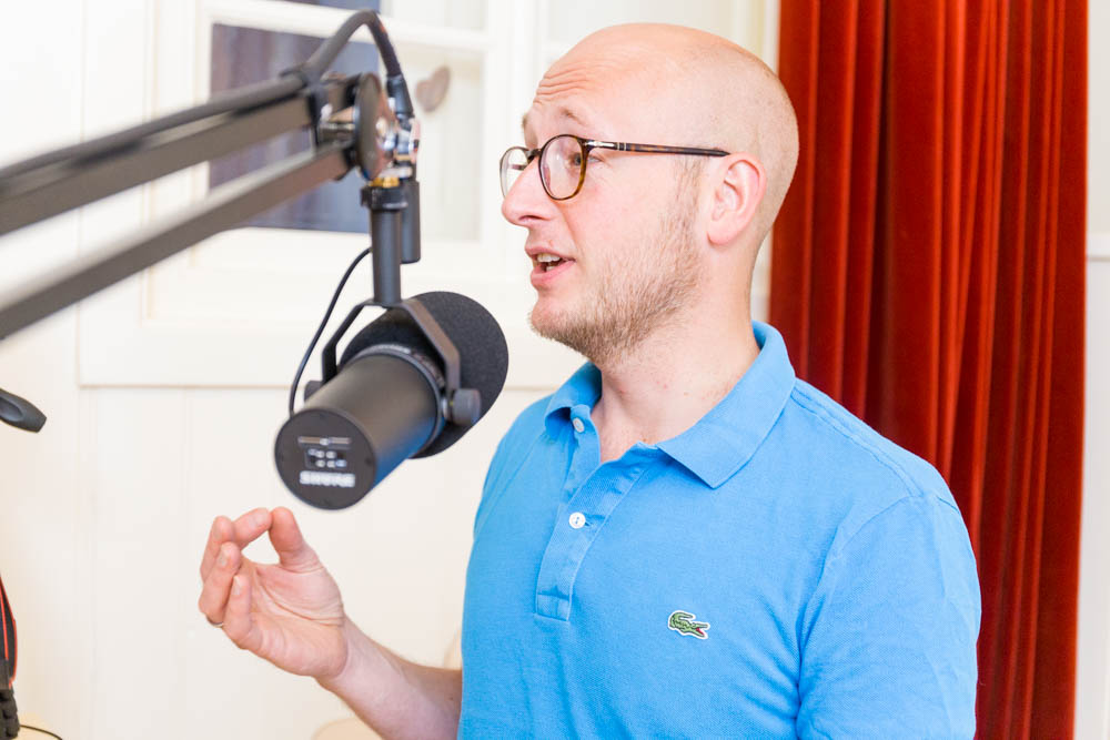 Man in blauwe polo spreekt in een microfoon tijdens een podcastopname.