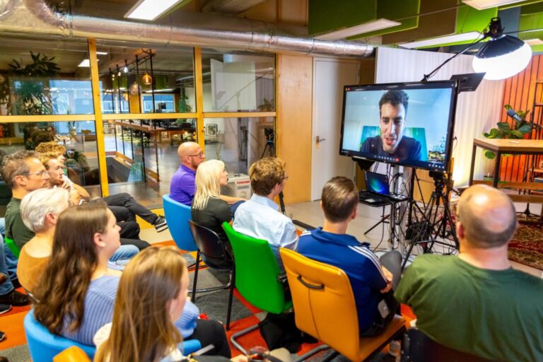 Een groep mensen kijkt naar een spreker op een groot videoscherm tijdens een All Ears Club-sessie met Lars Lensink.