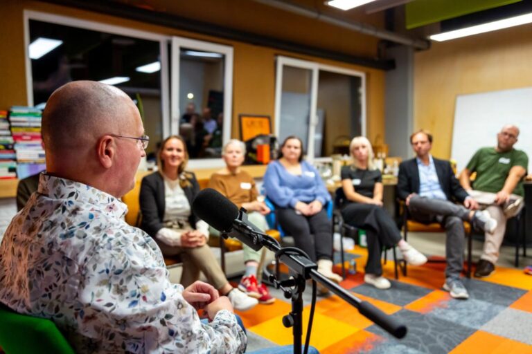 Man spreekt in microfoon tijdens een All Ears Club-sessie met aandachtig luisterend publiek in een kring.