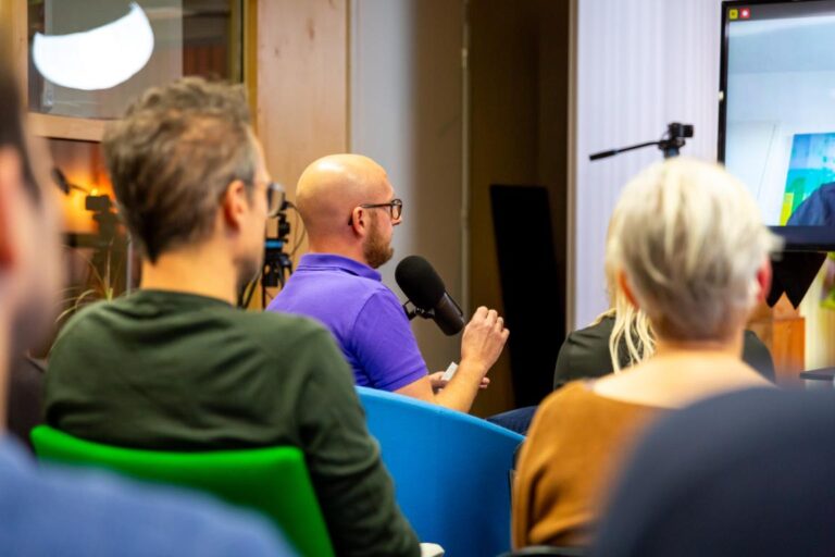 Een man in paarse polo stelt een vraag met een microfoon tijdens het All Ears Club-evenement met Lars Lensink.
