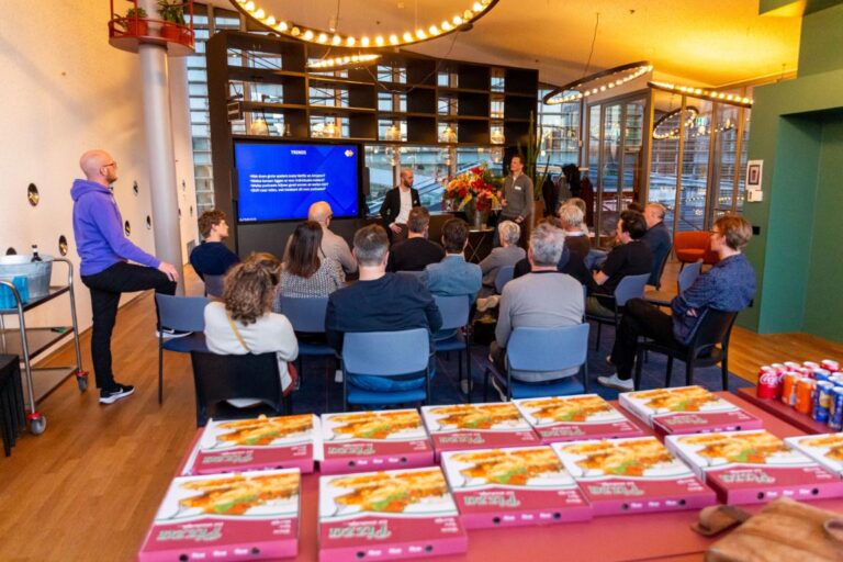 Een groep mensen woont een presentatie bij in een moderne ruimte bij NPO Luister, met dozen pizza en drankjes op de voorgrond.