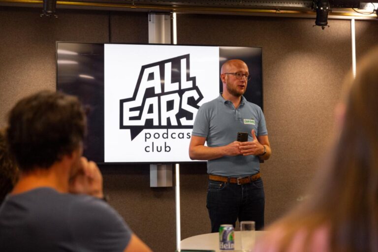 Man geeft presentatie bij het All Ears Podcast Club-event, met logo op scherm op de achtergrond.