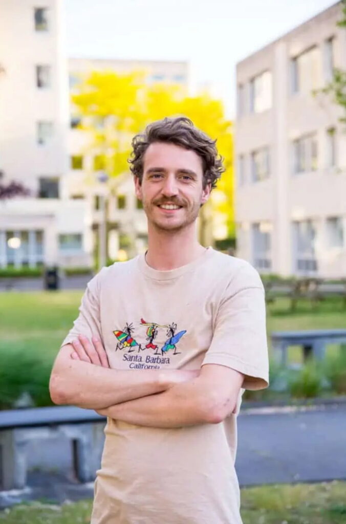 Man met opgerolde mouwen en een Santa Barbara T-shirt poseert buiten voor gebouwen en groen.