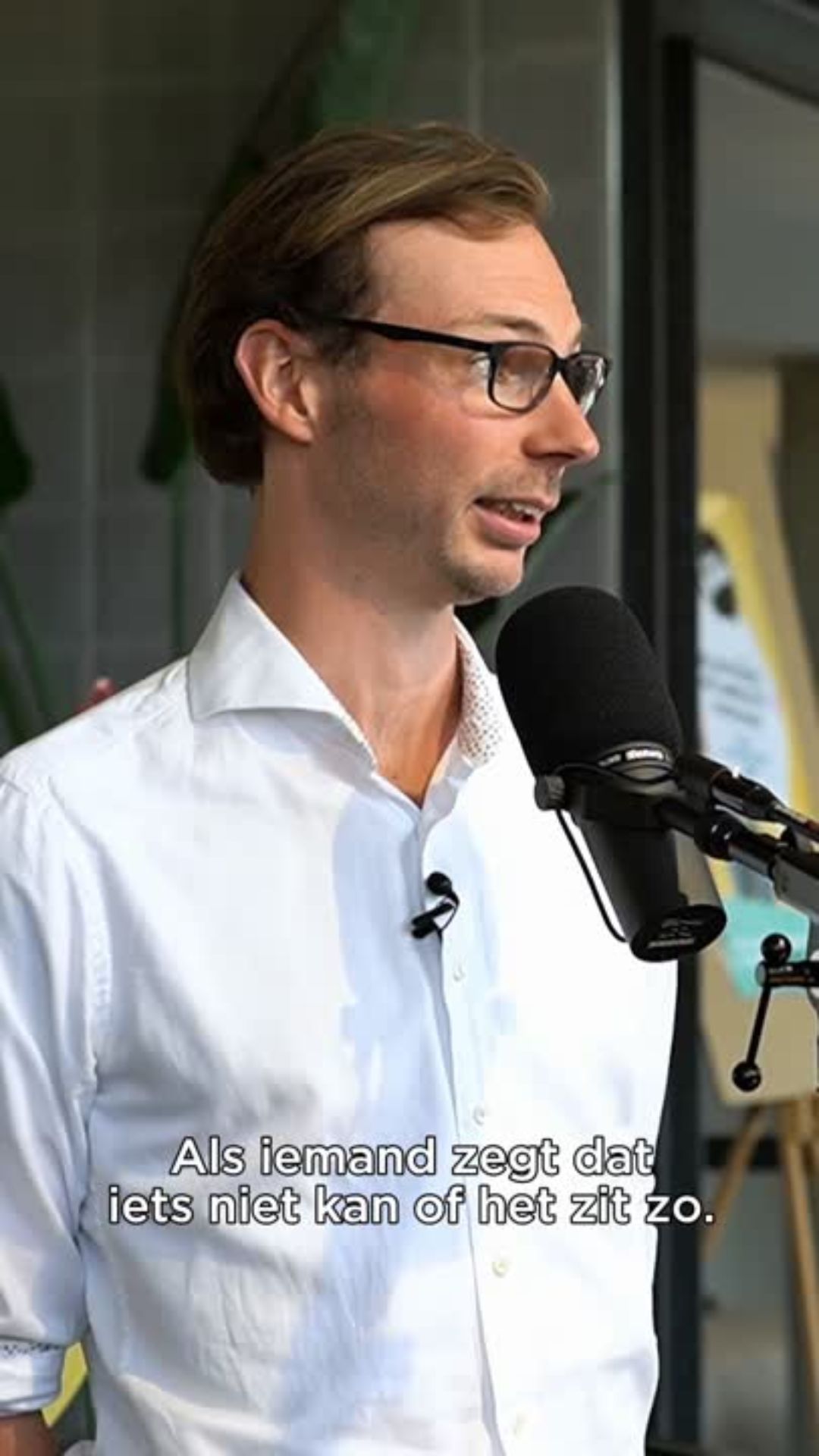 Man in een witte blouse spreekt in een microfoon tijdens een opname, met ondertiteling in beeld.