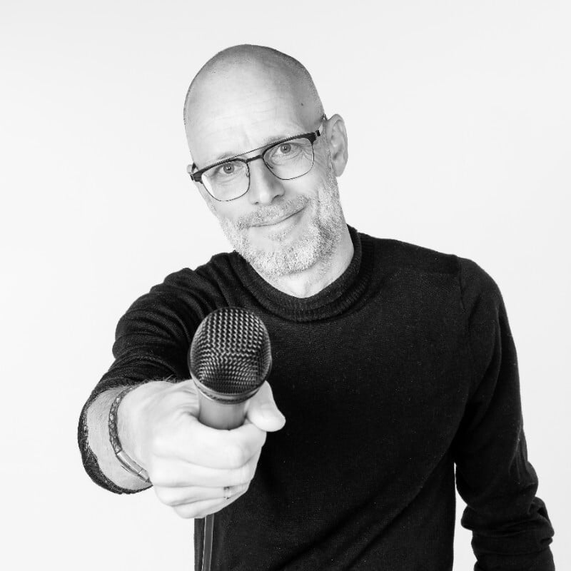 Man met bril en baard houdt glimlachend een microfoon naar de camera in zwart-wit portret.