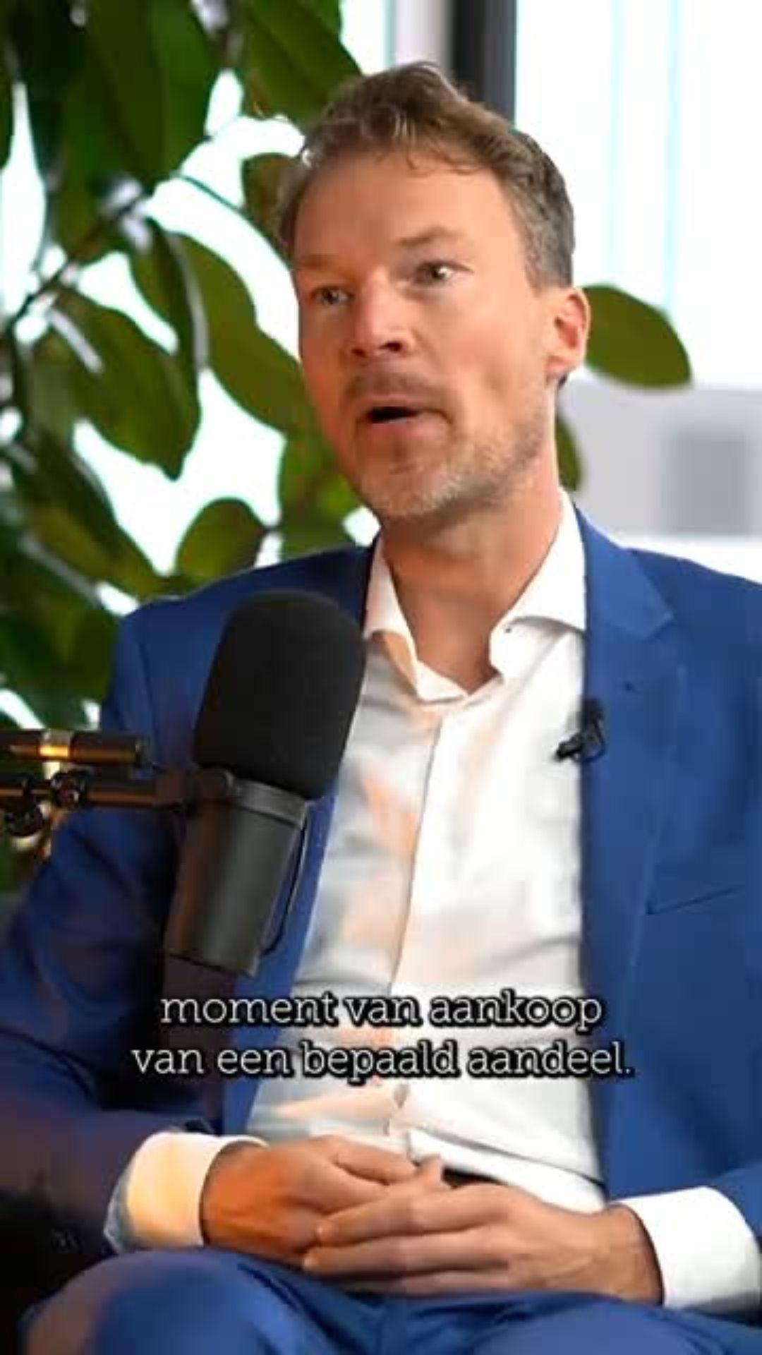 Man in blauw pak spreekt in een microfoon tijdens een interview over aandelen.